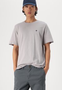Camiseta gris claro de manga corta con un logo sutil en el pecho, combinada con pantalones gris oscuro. Ambas prendas ofrecen un ajuste casual y relajado.
