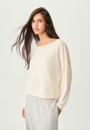 Helles beiges Longsleeve-Pullover mit quadratischem Ausschnitt und lässiger Passform, kombiniert mit grauen Sweatpants. Weicher Stoff, minimales Design.