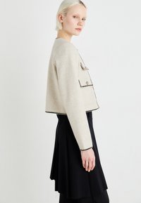 Crèmekleurig cropped jasje van zacht materiaal, met zwarte trim, twee voorkruppen en gouden knopen. Gecombineerd met een zwarte rok.