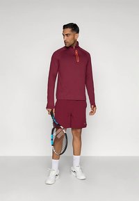 Burgundi sportöltözetet viselő férfi, aki teniszüstöt tart a kezében, egy sima fehér háttér előtt áll, oldalra nézve.