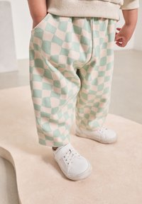 Hose aus weichem Stoff mit einem grün-beigen Schachbrettmuster, ausgestattet mit Seitentaschen und einer lockeren Passform, kombiniert mit weißen Turnschuhen.