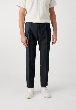 Zwarte broek gemaakt van gladde stof, slim fit met voorplooien, gecombineerd met witte sneakers en een getextureerd overhemd met korte mouwen.