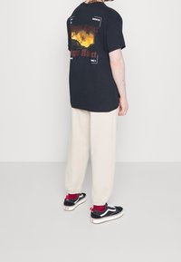 Camiseta gráfica negra con el estampado "Light Addict" en rojo en la parte posterior, combinada con pantalones de chándal crema y zapatillas negras con acentos rojos.
