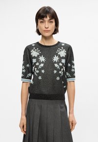 Μαύρο, κοντομάνικο τοπ με λαμπερό ύφασμα και λουλουδάτη κεντητική σε μπλε και λευκό. Ριμπ απόληξη στον λαιμό και τα μανίκια; εφαρμοστή γραμμή.