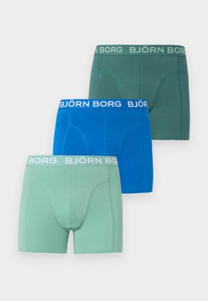 Homme torse nu debout portant un boxer Björn Borg bleu sur un sol recouvert de moquette claire avec un mur blanc uni en arrière-plan.