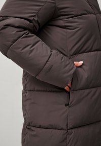 Parka in tessuto marrone scuro con design trapuntato. Presenta una tasca laterale e un'accents con logo sulla manica. Texture liscia in tutto il capo.