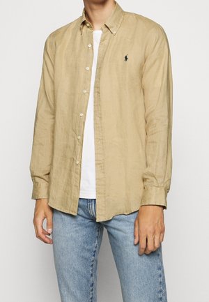 Man som bär en beige långärmad skjorta med knappar över en vit t-shirt och ljusblå jeans, stående mot en neutral bakgrund.