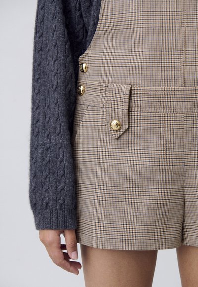 Pantalones cortos de cuadros marrones hechos de tela tejida, con botones dorados y un bolsillo de utilidades, combinados con un suéter de punto trenzado gris oscuro.