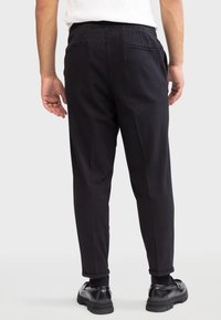 Pantalon noir avec une finition texturée, doté de poches arrière et d'une coupe fuselée. Porté avec des chaussures noires et des revers de cheville visibles.