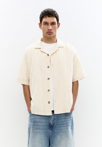Short-sleeve, light beige button-up shirt with a textured pattern, featuring dark button accents, paired with relaxed blue denim shorts.