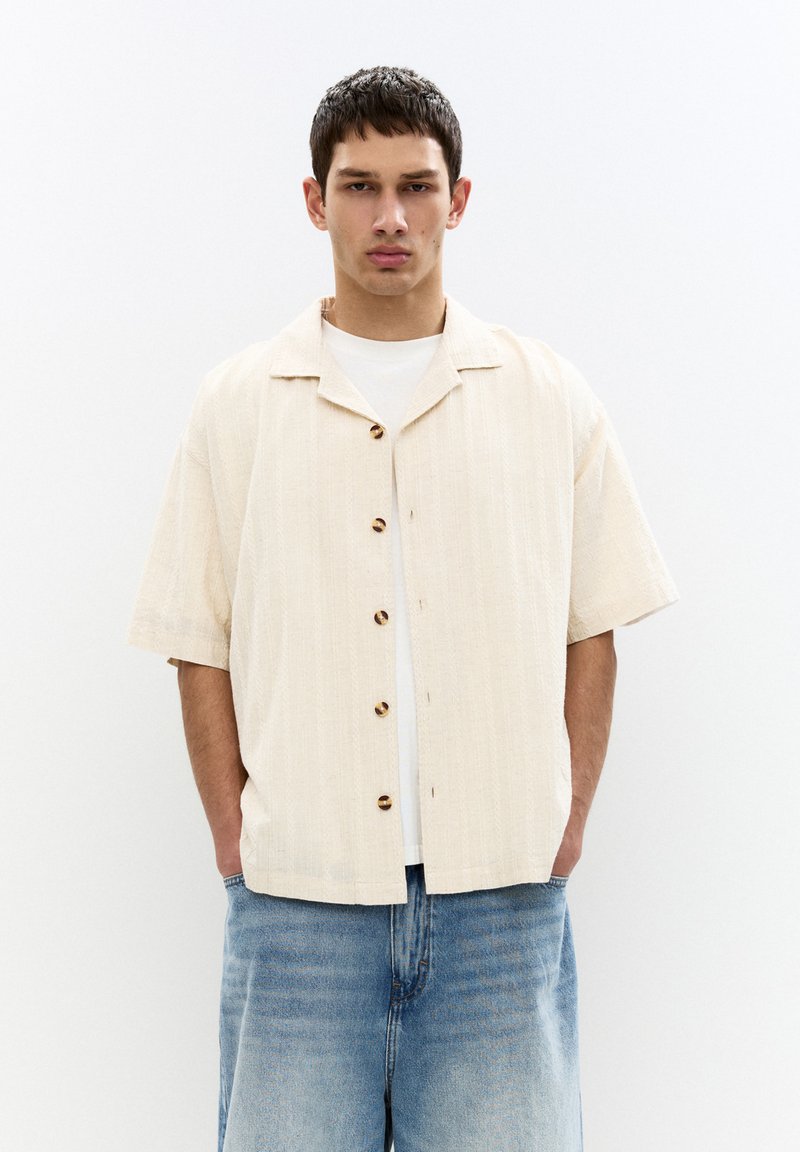 Short-sleeve, light beige button-up shirt with a textured pattern, featuring dark button accents, paired with relaxed blue denim shorts.