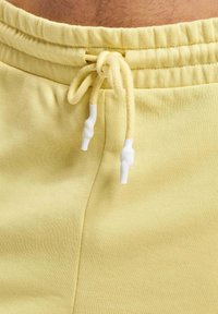 Gelbe Baumwoll-Sweatpants mit elastischem Bund und Kordelzug. Mit weißen Plastikenden am Kordelzug und glatter Textur.