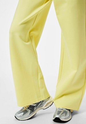 Jalad, mis kannavad lahtisi kollaseid pükse ja hõbedasi spordijalatsid, seisavad heledal taustal.