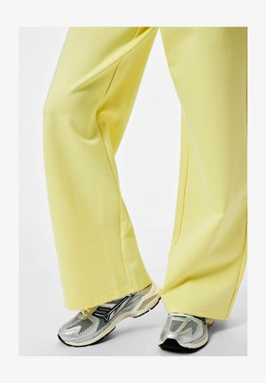 Jambes portant un pantalon jaune ample et des baskets argentées sportives, debout sur un fond clair.