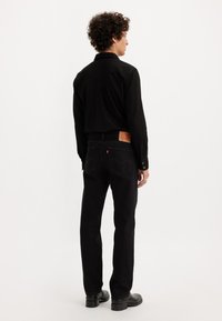 Moški s kodrastimi lasmi, oblečen v črn dolg rokav, črne kavbojke z usnjenim žepom in črne čevlje, stoji obrnjeno stran od bele ozadje.