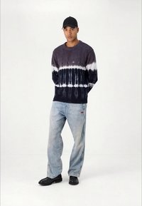 Knit sweater with dark blue and white tie-dye pattern, paired with light blue jeans and black footwear. Structured cap completes the outfit.