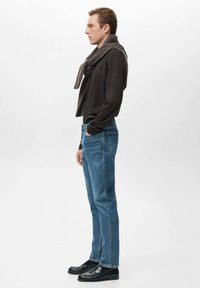 Camisa de manga larga oscura, vaqueros azules y mocasines negros. Una bufanda gris de punto cuelga alrededor del cuello. Perfil lateral contra un fondo claro.