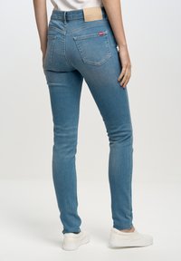 Gai zili džinsi ar šauru piegriezumu, pieci kabatas un brūns ādas jostas birka ar zīmola nosaukumu. Valkāti ar baltām slip-on kurpēm.