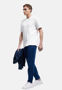 Weißes Baumwoll-T-Shirt, dunkelblaue Jeans mit strukturiertem Finish, hält eine dunkelblaue Jeansjacke, weiße Sneaker, steht vor einem schlichten Hintergrund.