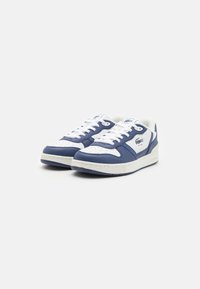 Zapatillas azul y blancas con materiales de cuero y textil, que cuentan con una caja de dedos perforada, diseño de cordones y suela de goma. Marca Lacoste visible.