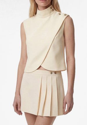 Frau trägt ärmelloses, beiges Wickel-Top mit Knopfdetails an Schulter und Kragen, kombiniert mit einem passenden plissierten Rock mit Knöpfen.
