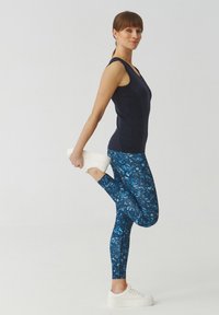 Marineblauw tanktop gecombineerd met blauwe leggings met abstracte spetters; model strekt haar been terwijl ze witte sportschoenen draagt.