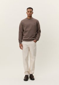 Sweatshirt marron à col rond, tissu léger, poignets côtelés, petit détail de logo. Associé à un pantalon beige clair et des chaussures marron foncé.