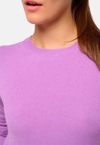 Gros plan sur une personne portant un pull en tricot violet clair avec un col rond, montrant une partie du visage et de longs cheveux bruns.