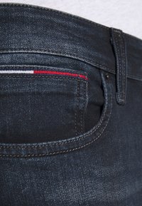 Jeans de mezclilla oscura con un primer plano del bolsillo trasero. Presenta detalles de costura en rojo, blanco y azul a lo largo del borde del bolsillo.
