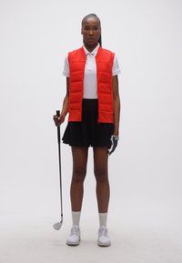 Γυναίκα με κόκκινο γιλέκο, λευκό πόλο, μαύρη φούστα και γάντι γκολφ που κρατάει μπαστούνι γκολφ, στέκεται μπροστά σε απλό φόντο.