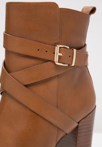 Hnědá kožená kotníková bota se zkříženým páskem a zlatou sponou, s blokovým podpatkem a hladkou texturou, se mírně špičatou špičkou.