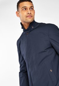 Trench coat azul marino con cuello alto, cierre de botón único y detalles de bolsillo lateral. Textura suave, diseño moderno.