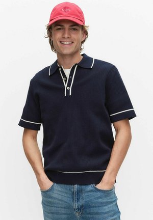 Jonge man in een marineblauwe korte mouw polo met witte bies en een rode pet, glimlachend met handen in de zakken van zijn blauwe spijkerbroek tegen een witte achtergrond.