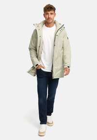 Lys grønn isolert jakke med høy krage, glidelås, elastiske mansjetter og lommer, kombinert med mørke jeans og hvite sneakers.