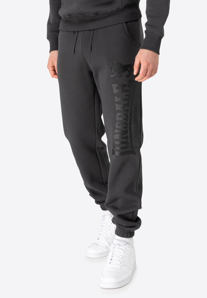 Pantalons de survêtement gris foncé en mélange de coton, avec un grand logo graphique noir sur la jambe, des poignets élastiques et des poches latérales. Portés avec des baskets blanches.