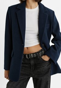 Marineblauer Blazer aus Wollmischgewebe mit Reverskragen, Ein-Knopf-Verschluss und zwei seitlichen Taschen. Getragen über einem weißen Oberteil und kombiniert mit hoch taillierten dunklen Jeans.