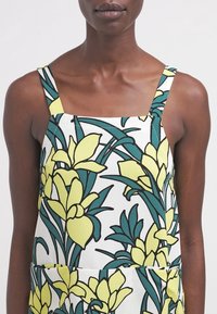 Blumenmuster-Kleidungsstück mit gelben und grünen Blumen auf weißem Hintergrund. Verfügt über breite Träger und einen quadratischen Ausschnitt. Glatte Stofftextur.