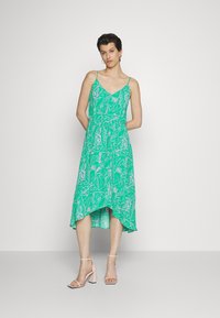 Vestido verde com alças finas, apresentando um padrão floral em tons mais claros. Dobra assimétrica na bainha, feito de um tecido leve e fluido.