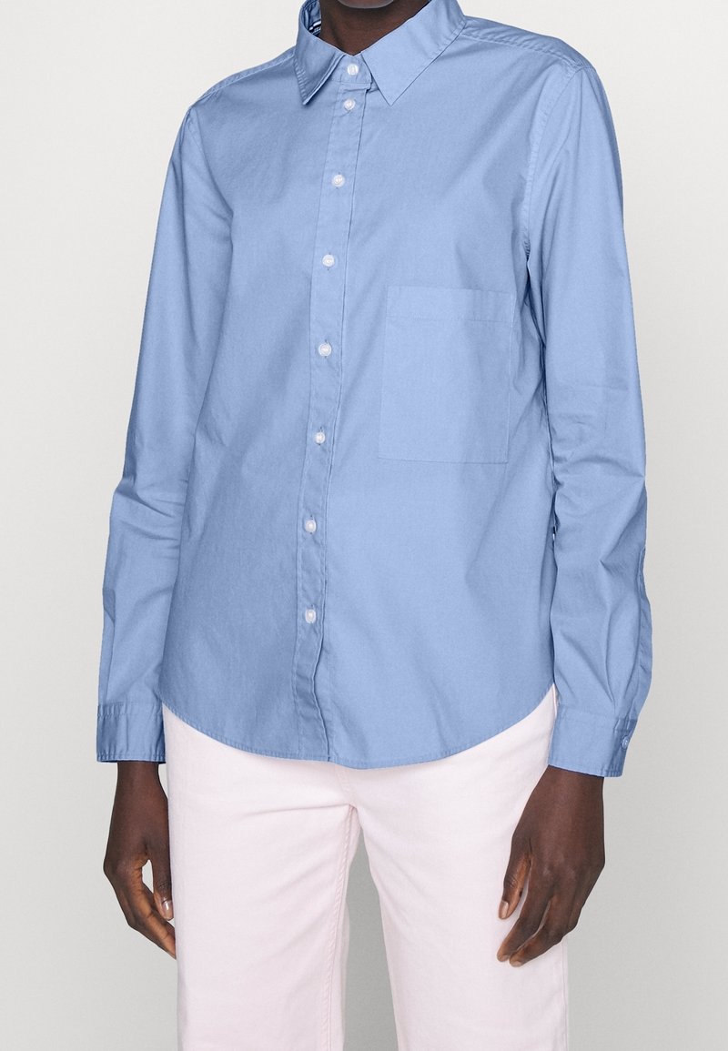 Ljusblå skjorta med knappar och spetsig krage, långa ärmar och bröstficka; tillverkad av ett mjukt tyg med en avslappnad passform.