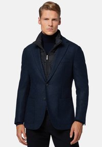 Blazer blu navy in tessuto di lana testurizzata, con due tasche frontali e un gilet in nylon nero imbottito sovrapposto. Presenta un colletto a risvolto.