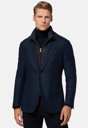 Navy blue blazer with a textured wool fabric, two front pockets, and a layered black quilted vest underneath. Features a notched collar.