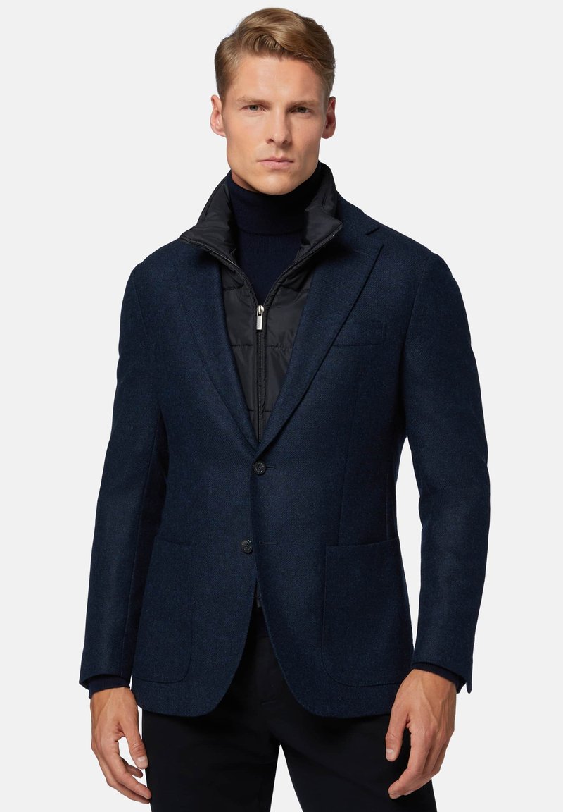 Blazer blu navy in tessuto di lana testurizzata, con due tasche frontali e un gilet in nylon nero imbottito sovrapposto. Presenta un colletto a risvolto.