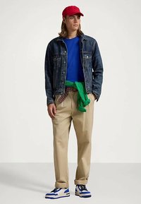 Ung person som bär en röd keps, jeansjacka, blå skjorta, beige byxor, grön tröja knuten i midjan och blå-och-vita sneakers.