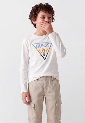 Un enfant souriant se couvre la bouche d’une main. Il porte un tee-shirt blanc avec le logo Guess et un pantalon cargo beige, sur un fond uni.