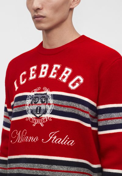 Gros plan d'une personne portant un pull rouge avec des rayures horizontales blanches, bleu marine et grises, texte « ICEBERG », blason et inscription « Milano Italia ».