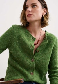 Cardigan vert avec un tricot texturé, encolure ronde et boutons en bois. Comprend des éclats de jaune et de bleu tout au long. Porté par-dessus un haut simple.