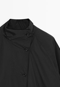 Zwarte asymmetrische gecoate jas met knopen, hoge kraag en diagonale naaddetails op een witte achtergrond.