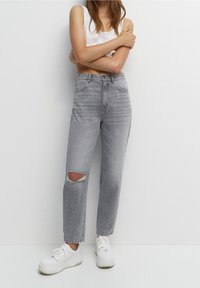 Jean gris taille haute avec une déchirure au niveau du genou, caractérisé par une coupe ample et une jambe légèrement fuselée, porté avec des baskets blanches et un débardeur blanc.