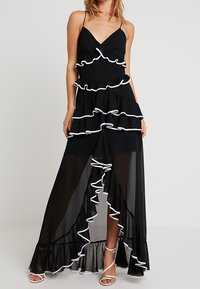 Schwarzes Maxikleid mit durchscheinenden Schichten, versehen mit weißen Rüschen entlang der Kanten und verstellbaren Spaghetti-Trägern. Weit schwingender Saum und entspannte Passform.