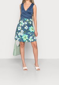 Blommig klänning med en marinblå bodice och V-ringning, samt en utsvängd kjol med gröna och vita blommor. Åtföljd av en mintgrön handv äska och ljusa klackar.